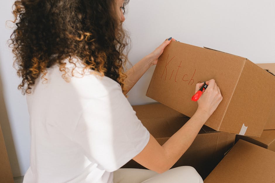 A woman with curly brown hair, facing away from the camera, is seen writing the word 'Kitchen' in red marker on the side of a small cardboard box. She is wearing a white t-shirt and is surrounded by s